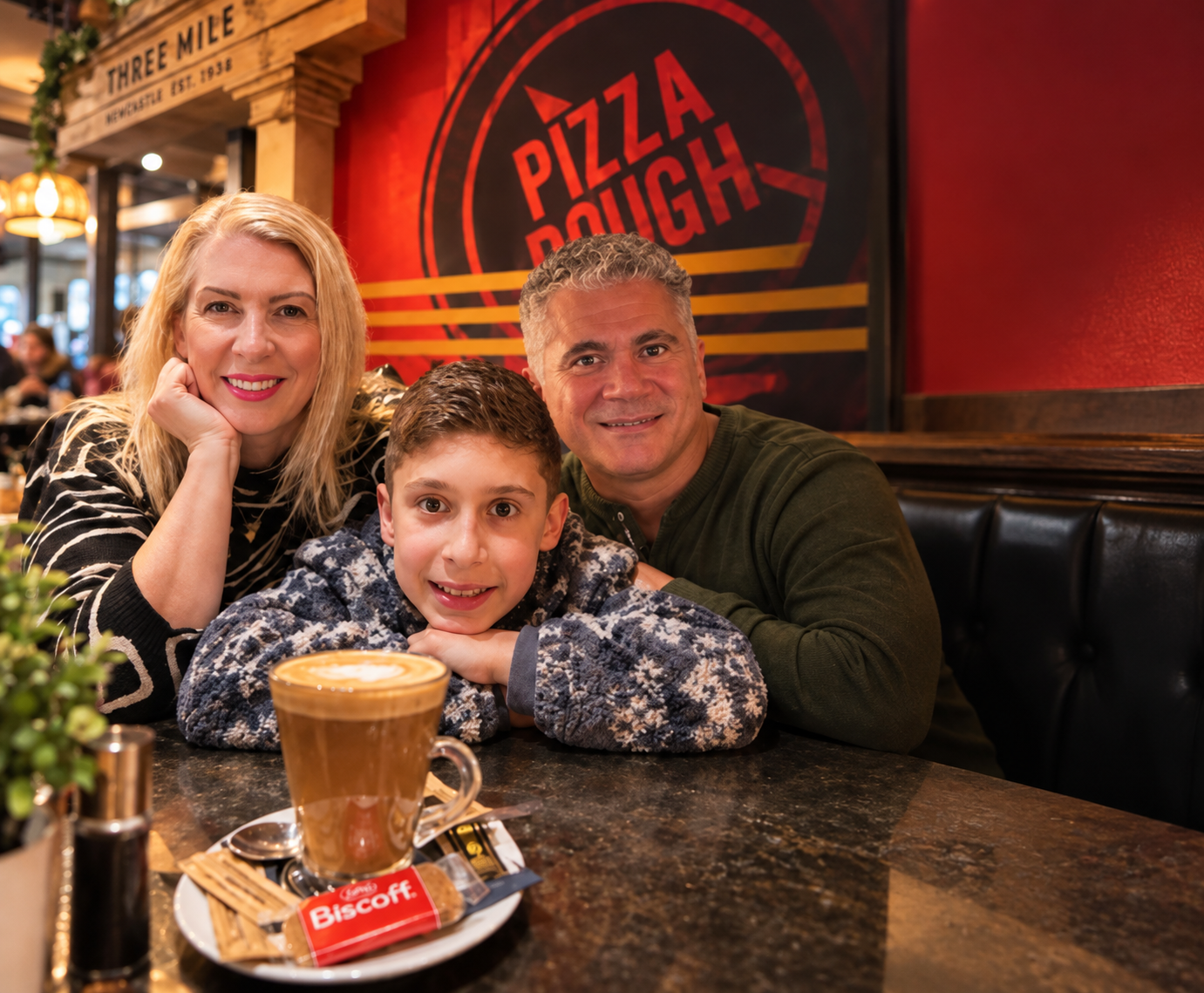 Saffron, H and Cyrus smiling together at a restaurant table in Newcastle, with a latte in the foreground and a Pizza Dough sign behind them.